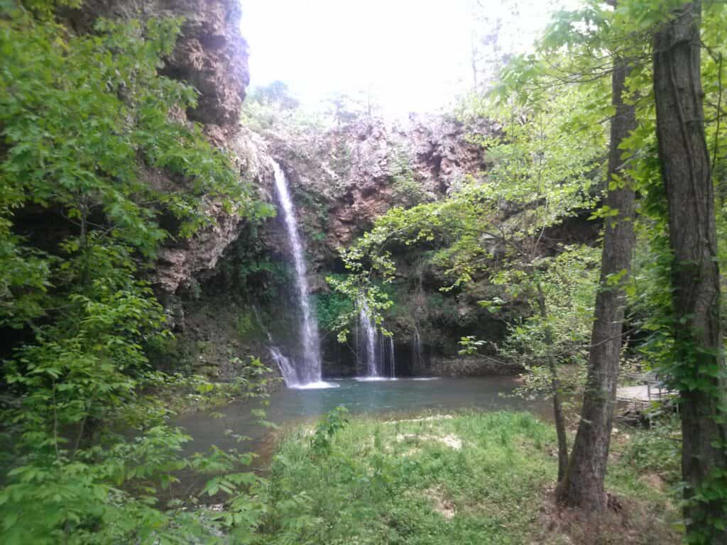 Dripping Springs Waterfall falls over a rocky cliffside in a narrow band of water into the pool below and is surrounded by the lush greenery of Natural Falls State Park, Oklahoma.