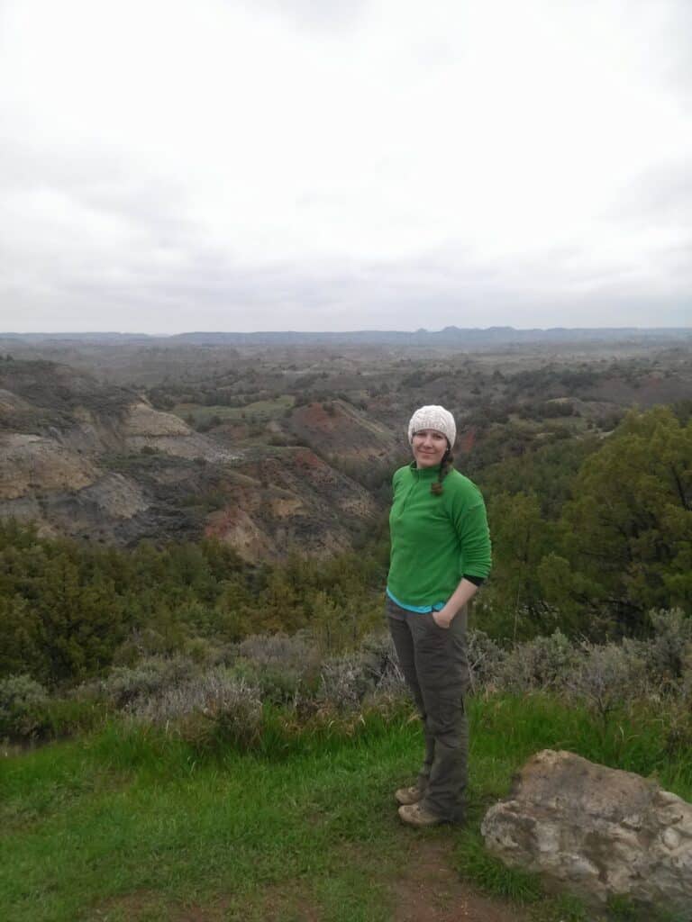 Woman in green sweater and brown hiking pants stands above a view of the badlands of the Theodore Roosevelt National Park South Unit.