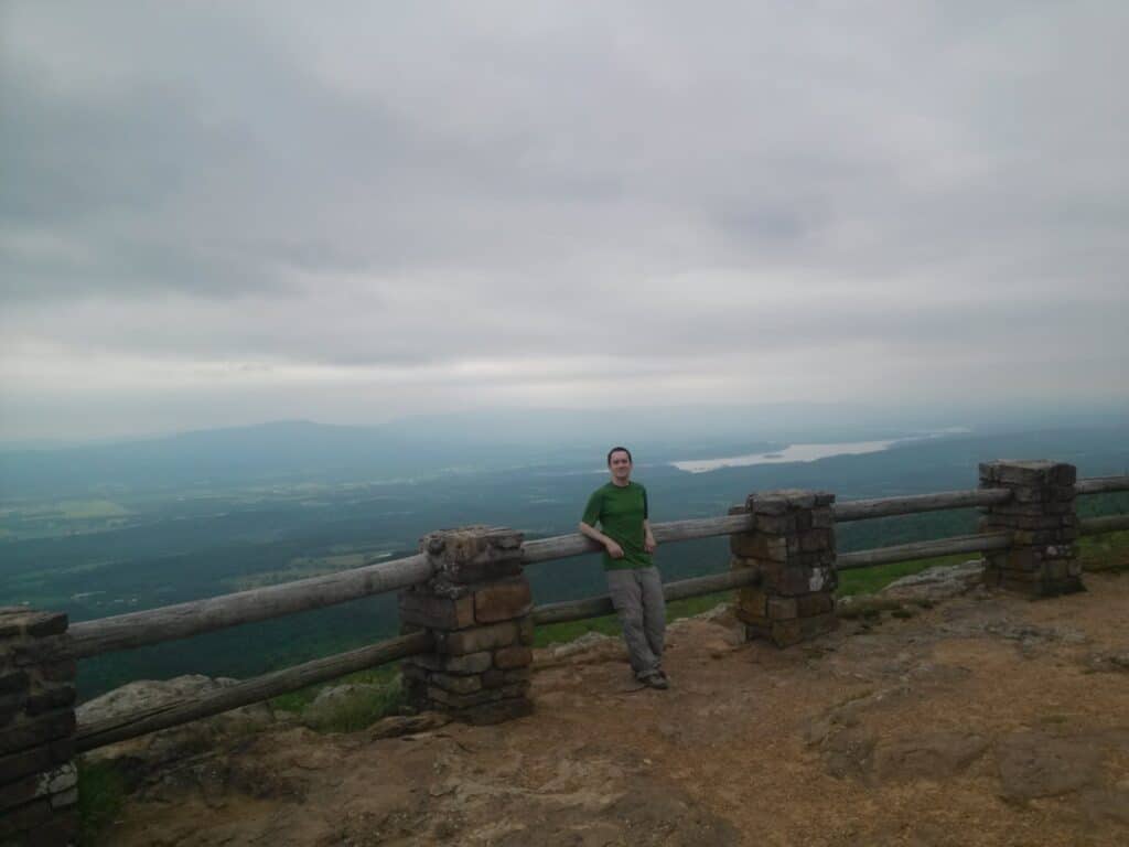 Scenic outlook at Mount Magazine State Park over a lush rolling landscape. A man leans against a wood and stone barrier facing the camera.