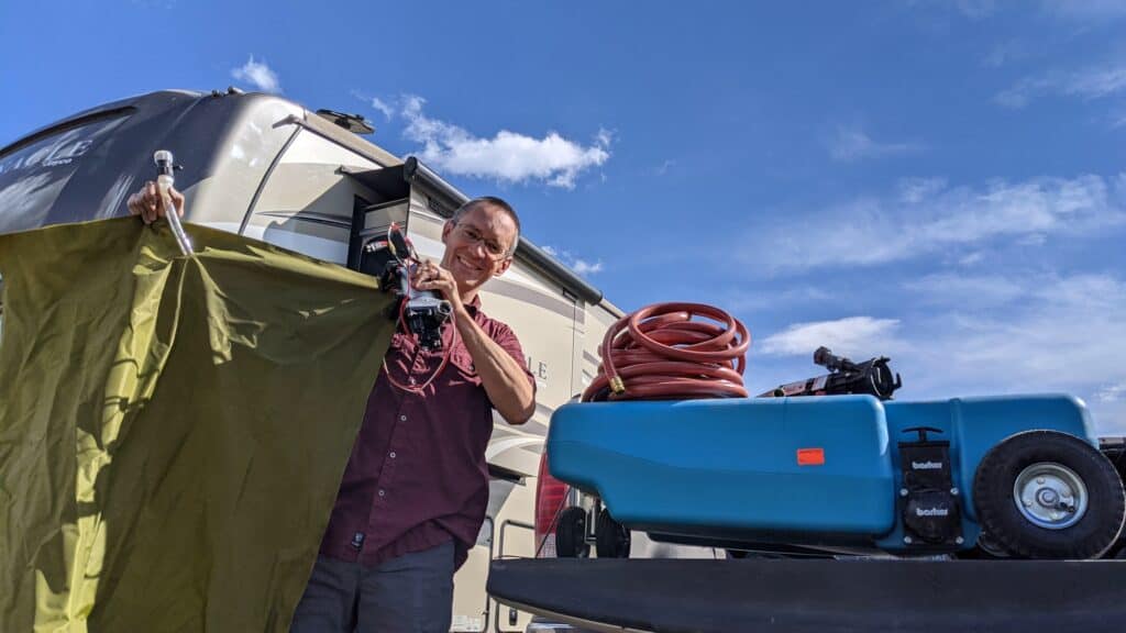 A man smiles while setting up RV waste equipment, holding a water bladder and pump beside a portable blue waste tank and coiled red hose. This behind-the-scenes view highlights one of the practical aspects of full-time RV living.