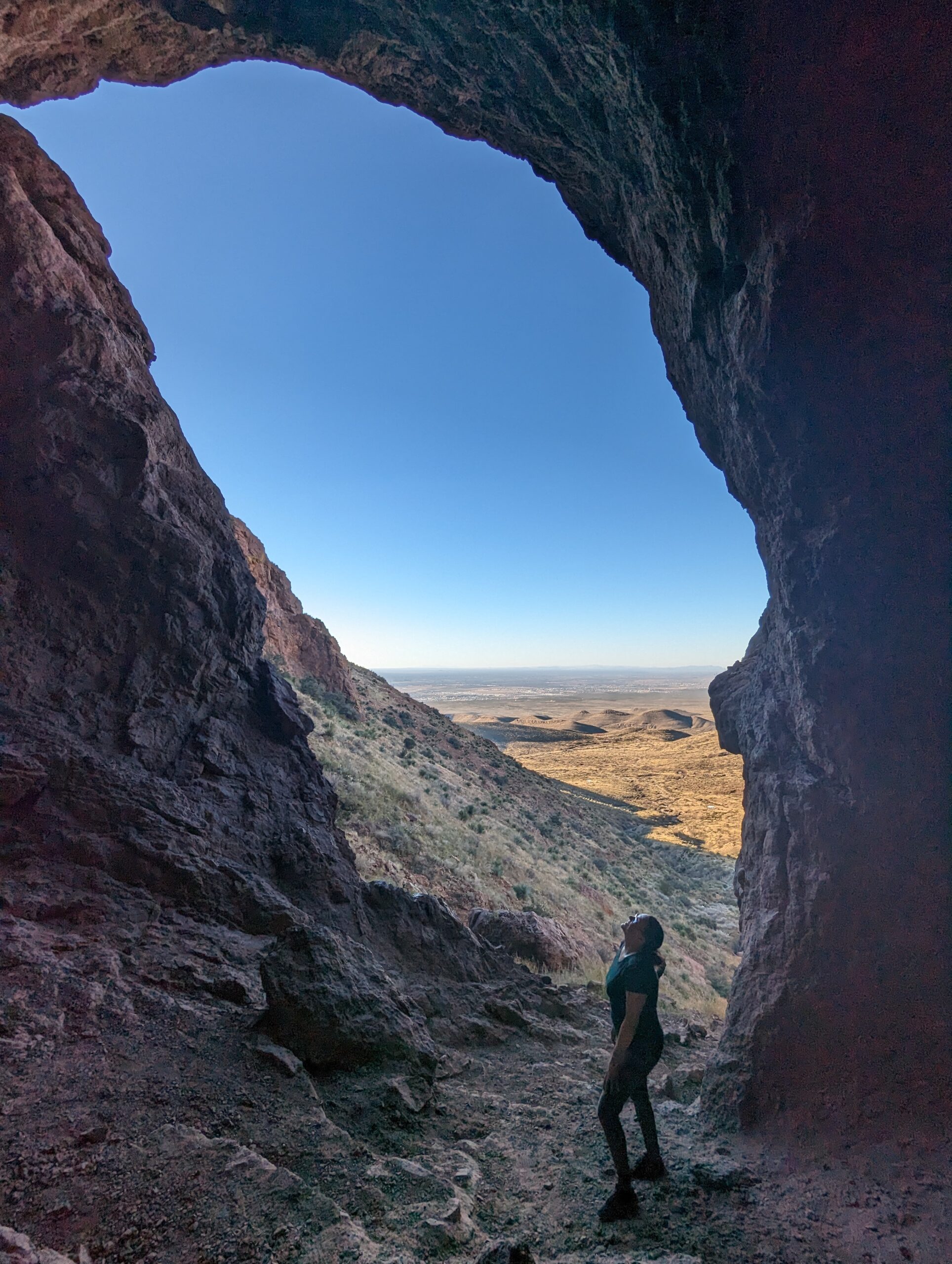 Woman stands in the oval entrance of the Aztec Caves in the Franklin Mountains of El Paso Texas, silhouetted by the landscape and blue sky behind her