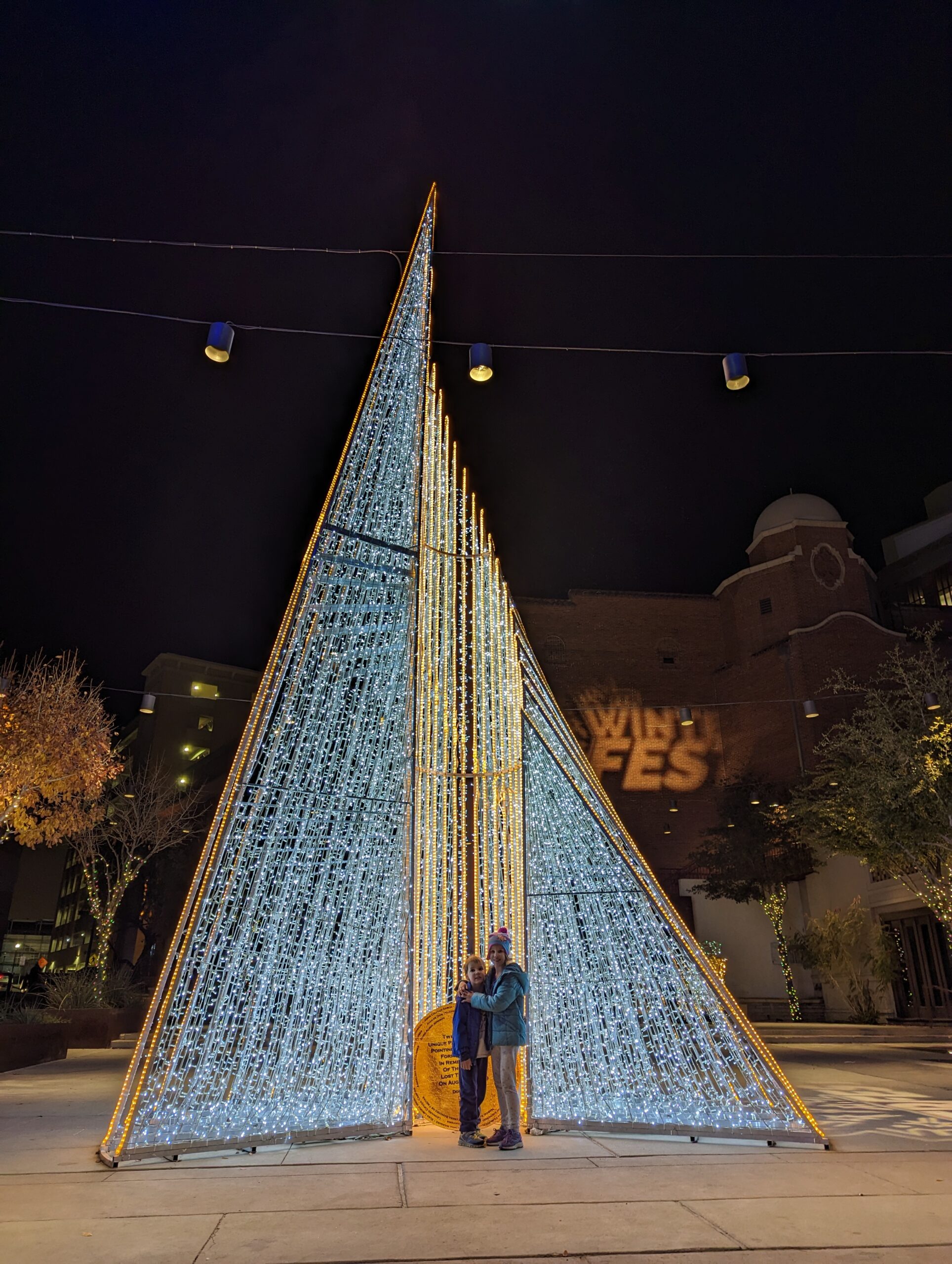 Two kids stand in front of a large triangular Christmas lights structure for Winter Fest in El Paso.