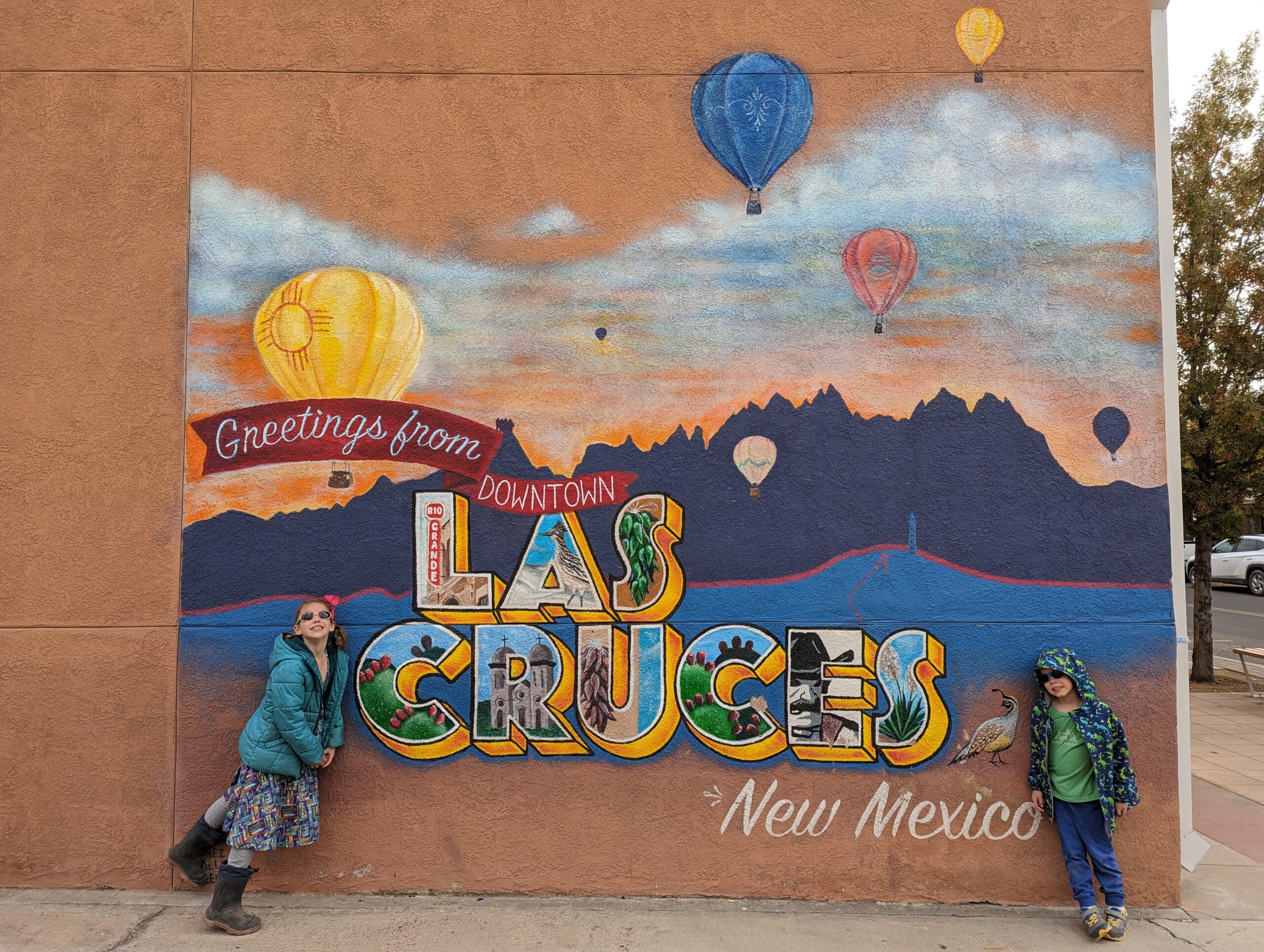 Two kids leaning on a wall with a mural with mountains, hot air balloons, and the text Greetings from Downtown Las Cruces New Mexico.