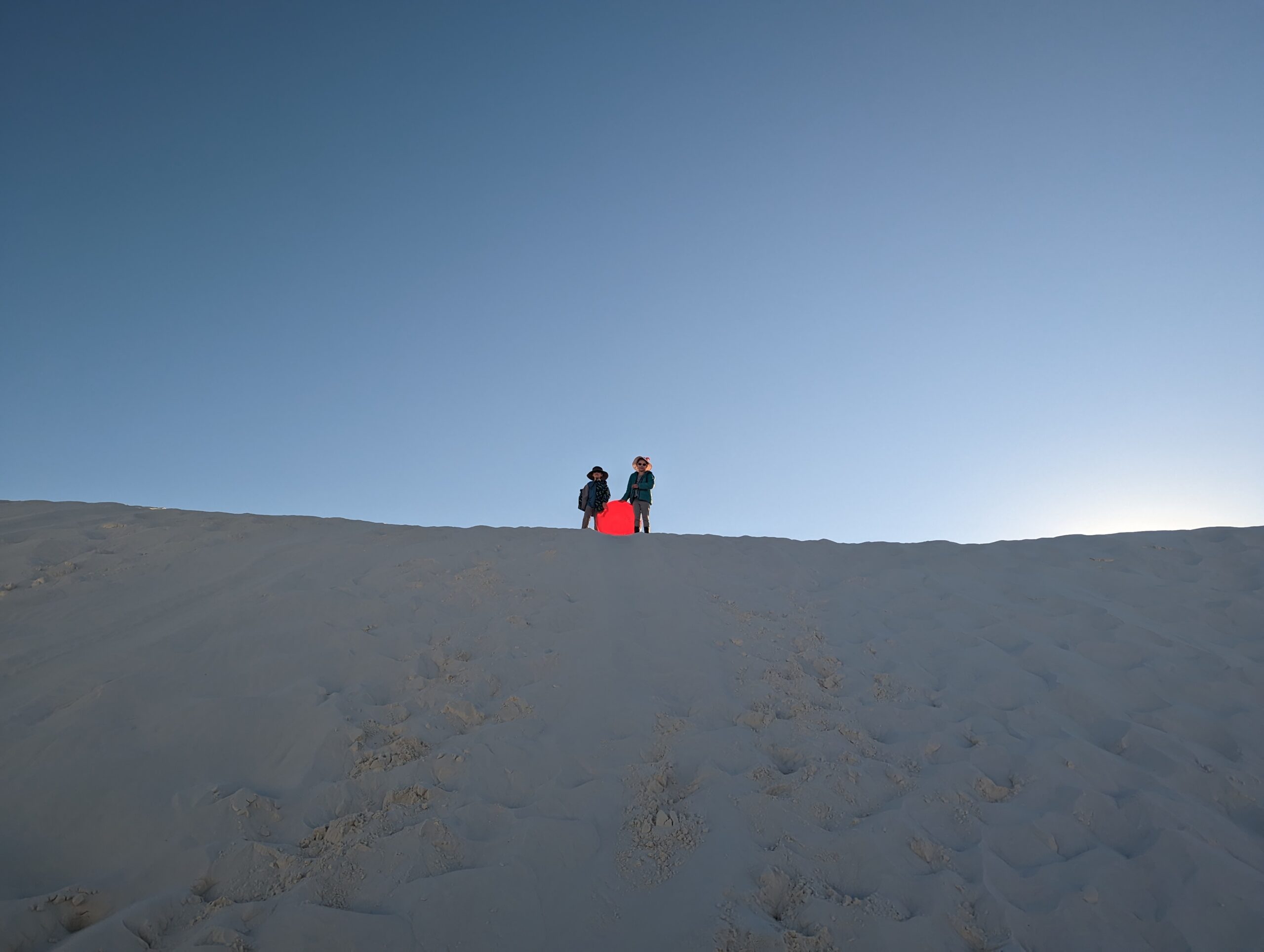 Two kids stand holding a round sled on the top of a dune at White Sands National Park.