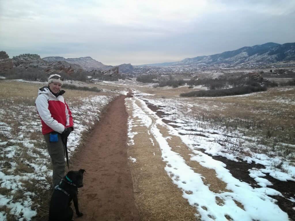 A woman in a white and red winter coat stands on a trail with her leashed black dog. The trail goes downhill through a snow covered landscape of red rock outcroppings at South Valley Park.