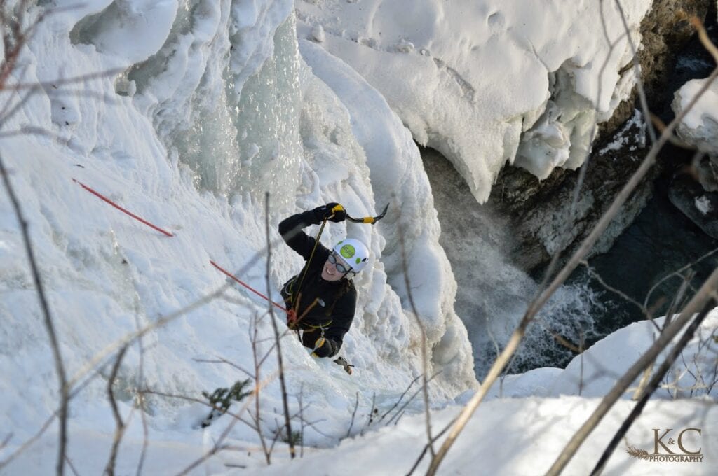 A woman as seen from above, is ice climbing on a frozen waterfall near Telluride. She is attached to a rope and swinging an ice axe toward the ice.