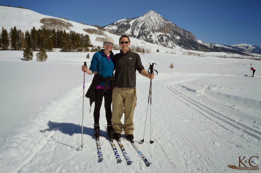 A couple standing together on a groomed cross-country skiing trail with poles in hand and Crested Butte Mountain behind them. 