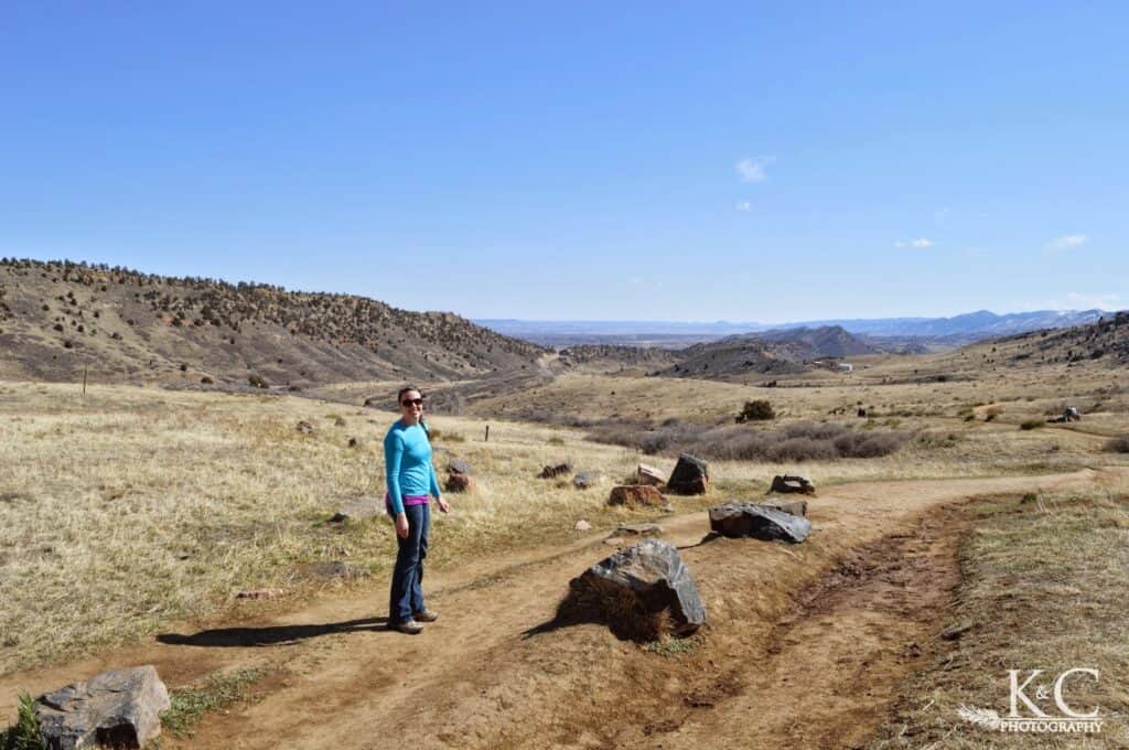 A woman stands at the beginning of a dirt hiking trail in Matthews / Winters Park near Denver, Colorado, surrounded by dry grassy hills and distant rocky ridges under a clear blue sky.