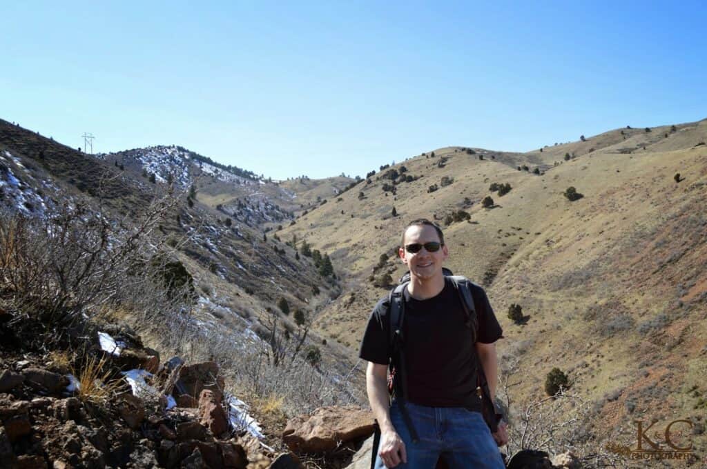 A man wearing sunglasses and a backpack leans against a rock on a hillside trail in Matthews / Winters Park near Denver, Colorado, with snowy patches and rugged hills stretching out behind him under a clear blue sky.
