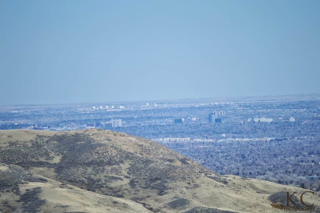 View from a high point in Matthews / Winters Park looking out toward the Denver metro area, with grassy hills in the foreground and city buildings stretching across the horizon under a hazy blue sky.