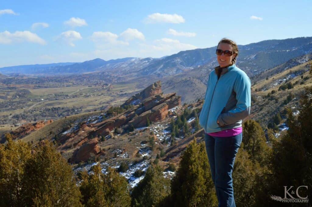 A woman in a blue jacket and sunglasses stands at a scenic overlook in Matthews / Winters Park with views of Red Rocks Park and Amphitheater red rock formations, snowy foothills, and the surrounding Colorado valley near Denver.