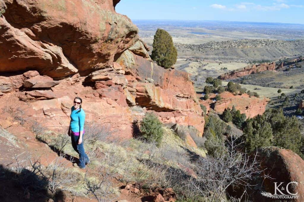 A woman stands beside large red rock formations along a trail in Matthews / Winters Park near Denver, Colorado. The scenic overlook reveals layered sandstone cliffs and sweeping views of the surrounding foothills and plains.