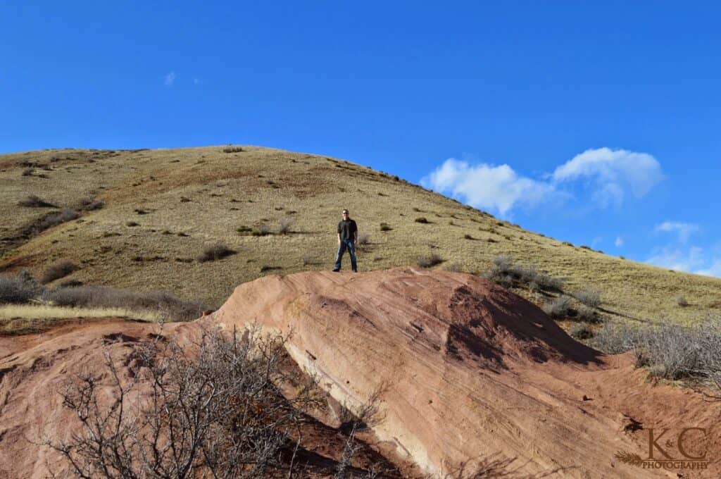A man stands on a large red sandstone formation at Matthews / Winters Park, with golden grassy hills rising behind him under a vivid blue sky. The striking landscape showcases the unique geology and hiking terrain in the park.