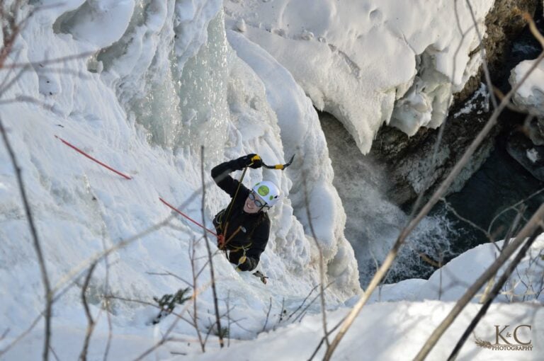 A woman as seen from above, is ice climbing on a frozen waterfall near Telluride. She is attached to a rope and swinging an ice axe toward the ice.