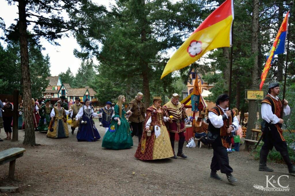 Colorado Renaissance Festival parade of characters dressed in period costume. They are walking in pairs of two, man and woman, with two men leading the way holding flags.