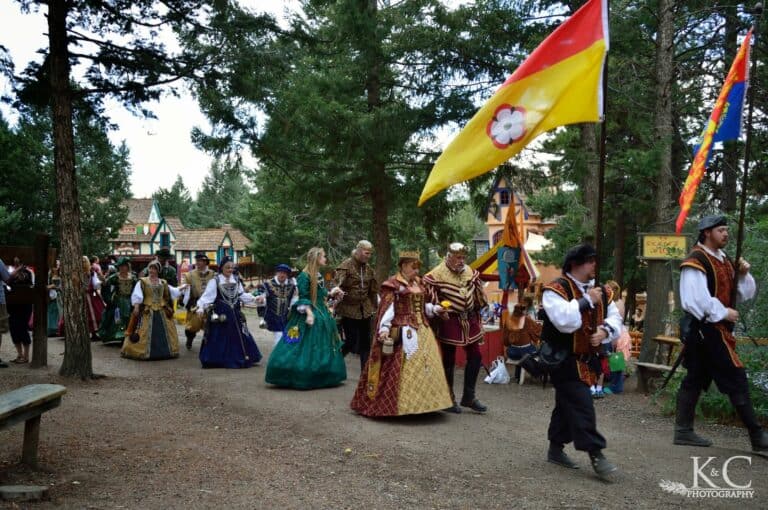 Colorado Renaissance Festival parade of characters dressed in period costume. They are walking in pairs of two, man and woman, with two men leading the way holding flags.