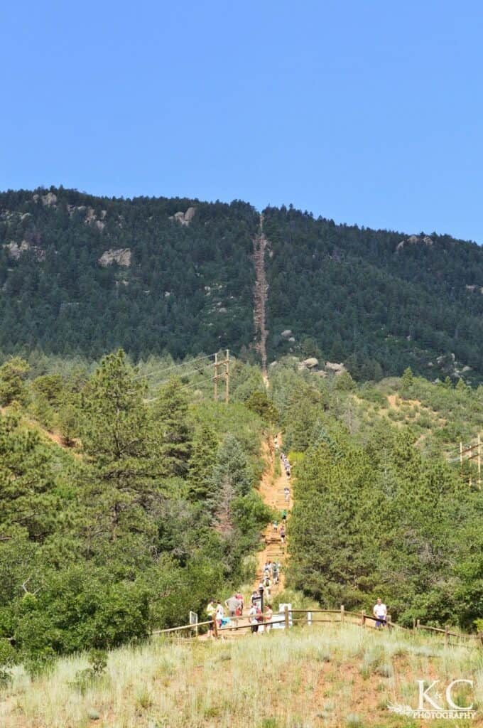 The Manitou Incline is seen from below with many stairs climbing straight up the mountain side with many people on its steps taking this strenuous hike.