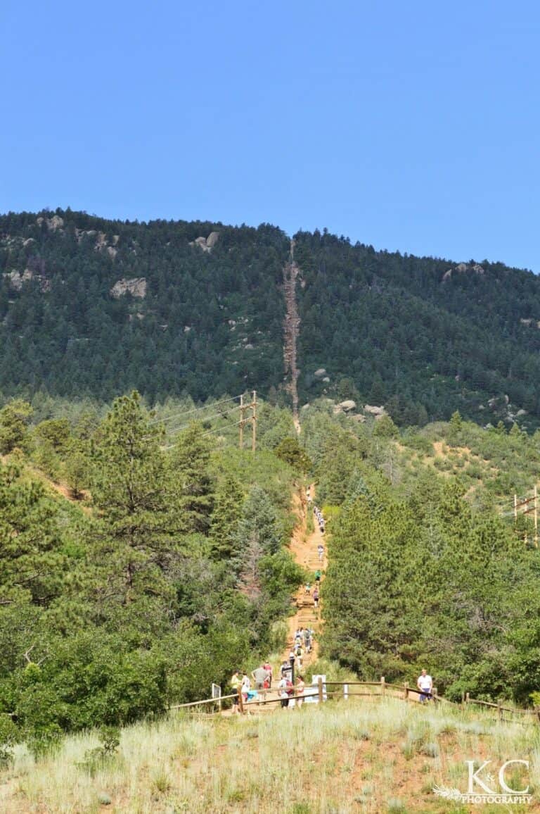 The Manitou Incline is seen from below with many stairs climbing straight up the mountain side with many people on its steps taking this strenuous hike.