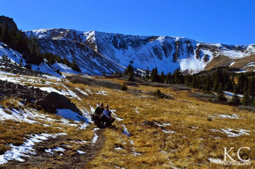 A man and woman crouch on the trail with their black dog near the top of Butler Gulch Trail with snow covered mountains behind.
