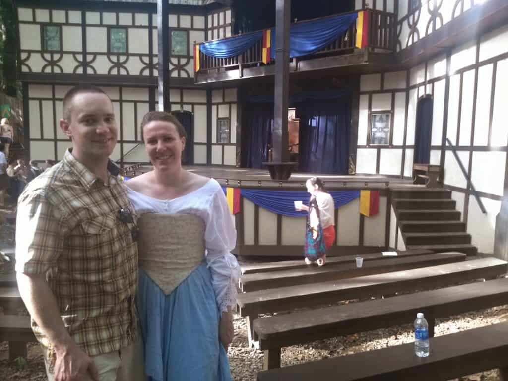 A man and woman stand in front of a medieval stage at the Maryland Renaissance Festival. She is dressed in period costume while he wears modern clothing.