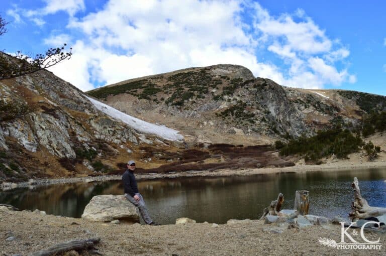 A man leans against a rock in front of the lake below St. Mary's Glacier. The snowfield is seen on the hillside above the lake.