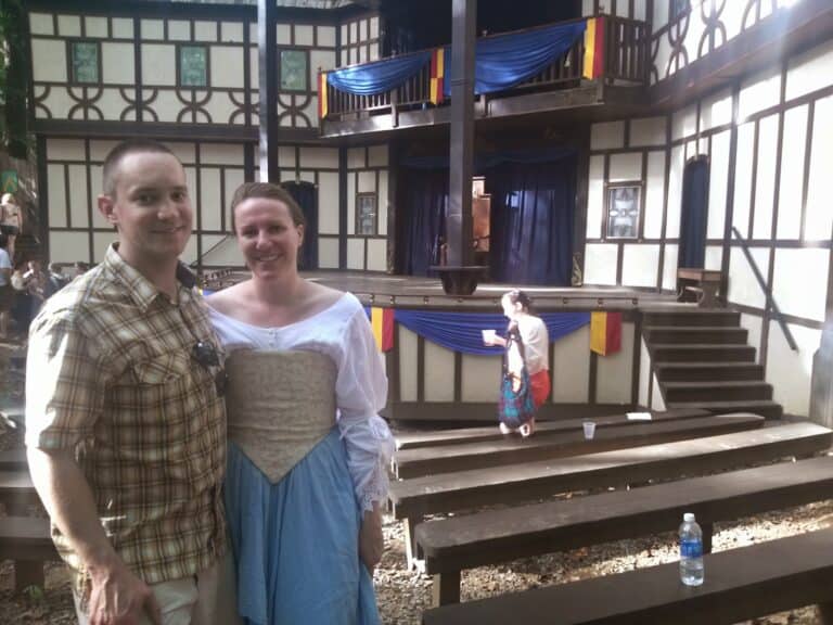 A man and woman stand in front of a medieval stage at the Maryland Renaissance Festival. She is dressed in period costume while he wears modern clothing.