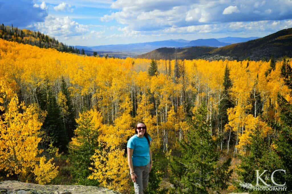 Woman standing in front of bright yellow aspen trees at an overlook. Steamboat Springs fall colors as far as you can see, with mountains in the distance.