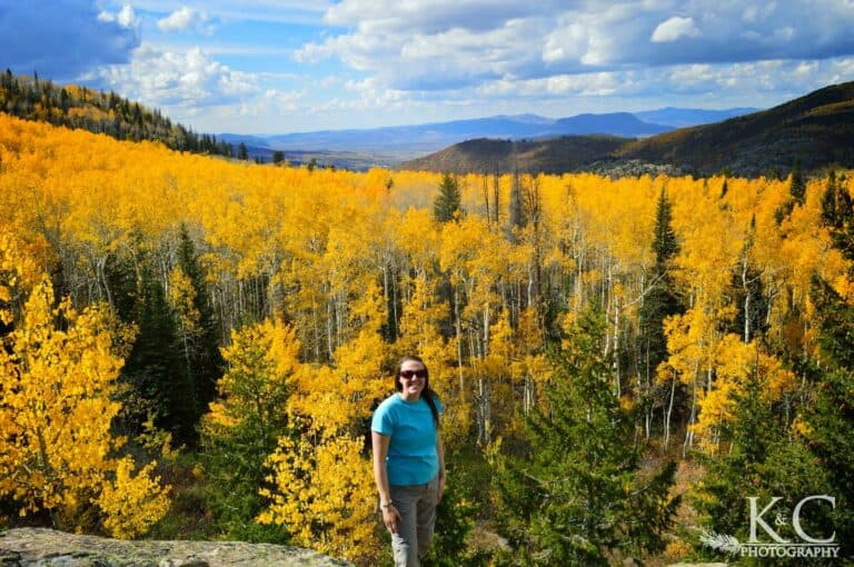 Woman standing in front of bright yellow aspen trees at an overlook. Steamboat Springs fall colors as far as you can see, with mountains in the distance.