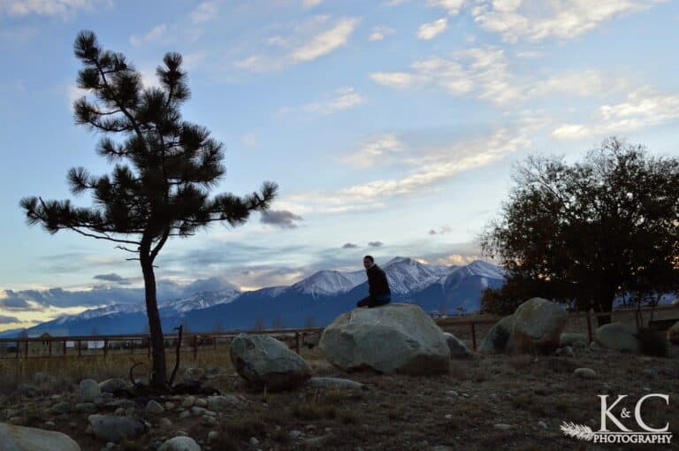 Woman sitting on top of a large rock looking over her shoulder in Buena Vista Colorado with the snow covered Collegiate Peaks in the distance.