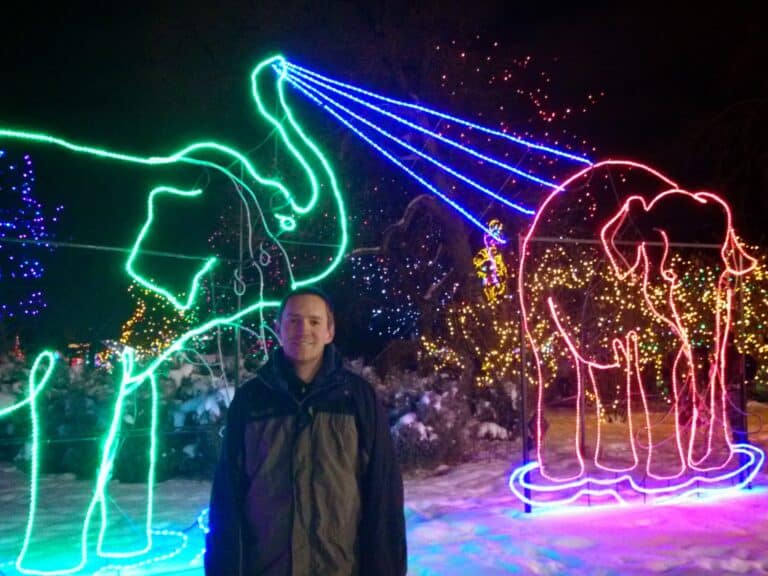 Man standing in the snow in front of the Denver Zoo Lights festive Christmas display showing a green elephant spraying water on a red elephant.