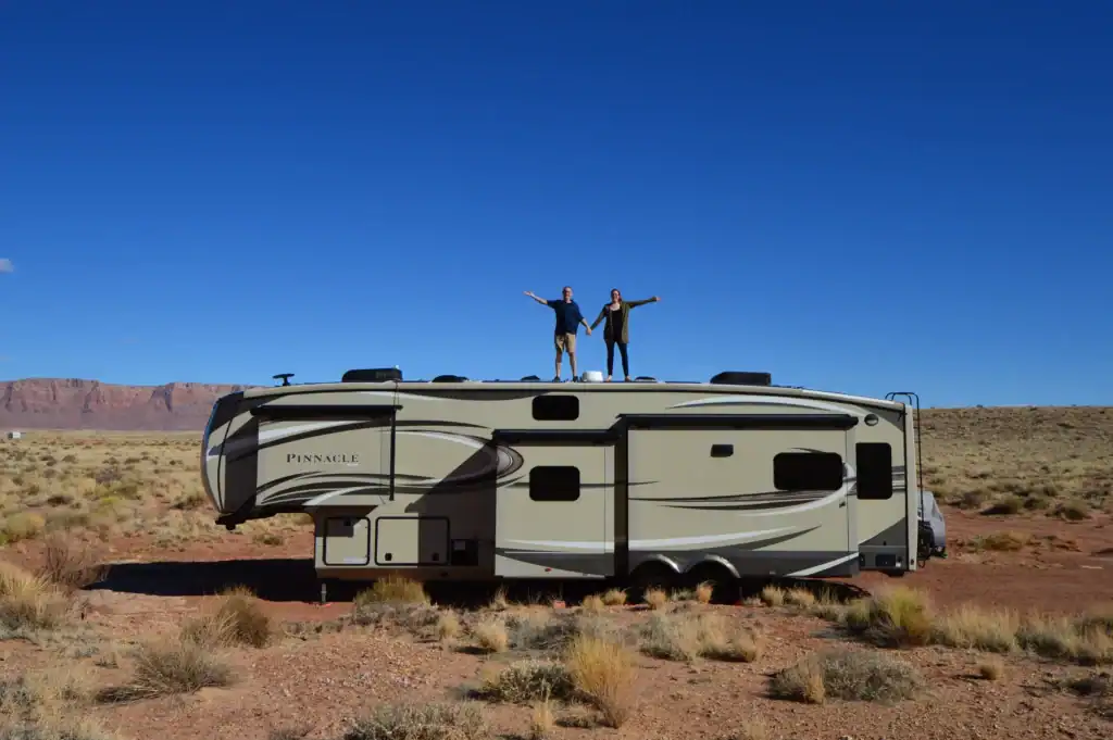 A couple stands on top of their large Jayco Pinnacle RV with arms outstretched under a clear blue sky, surrounded by desert landscape and red rock cliffs. This photo captures the adventurous spirit of their family travel blog and RV lifestyle.