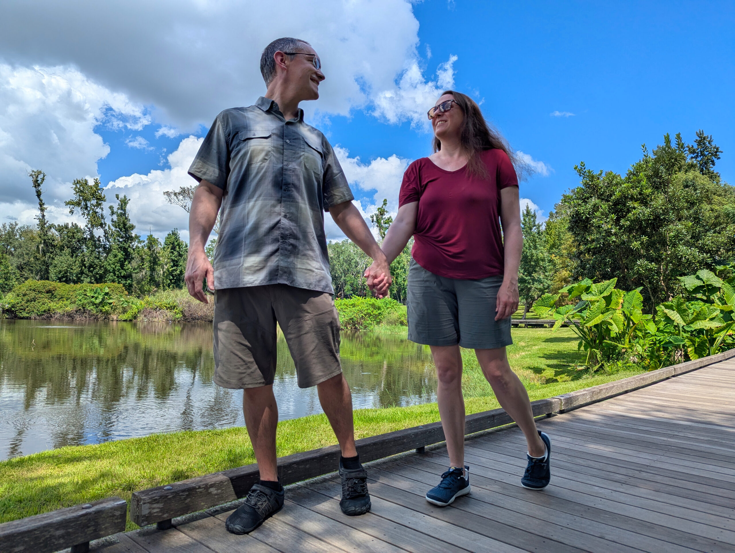 A couple walks hand in hand on a wooden walkway next to a pond and green trees, both wearing KUHL shorts. The scene showcases the shorts' relaxed fit and breathable fabric, perfect for comfortable strolls in warm weather.