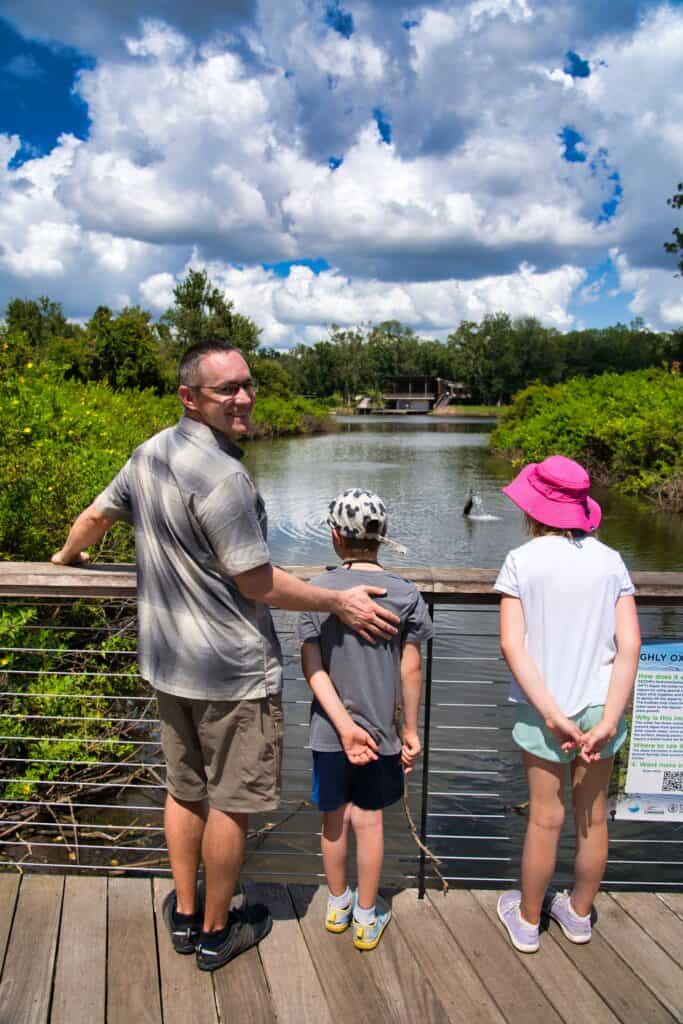 A man wearing KUHL shorts and a plaid shirt stands on a wooden boardwalk with two children, looking out over a calm lake under a bright, cloud-filled sky. The scene highlights the versatility of KUHL shorts for warm-weather exploration and relaxed family outings in nature.