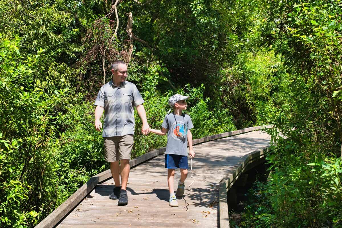 A man wearing KUHL shorts and a plaid hiking shirt walks hand-in-hand with a young boy on a wooden boardwalk trail surrounded by dense greenery. The sunny setting highlights the comfort and functionality of the KUHL shorts for outdoor adventures.