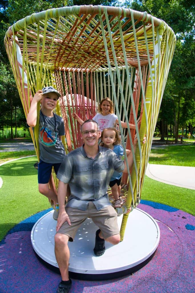 A man wearing tan KUHL shorts and a plaid shirt poses with his three smiling children inside a colorful woven playground structure on a sunny day. The shorts' casual fit and performance fabric make them ideal for active family outings and warm-weather adventures.