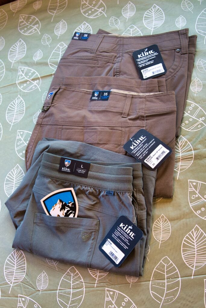 Three pairs of KUHL shorts in earthy tones are laid out on a green leaf-patterned background, with visible tags showing sizes and branding. The display highlights different styles and features, including a logo patch and stretchy waistband for comfort and versatility.