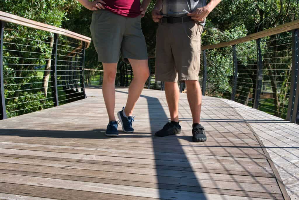 Two people standing on a wooden pedestrian bridge wearing KUHL shorts, with one in olive green women's shorts and the other in tan men's shorts, both styled for outdoor wear and casual comfort. Their legs and footwear are visible, with lush greenery in the background.
