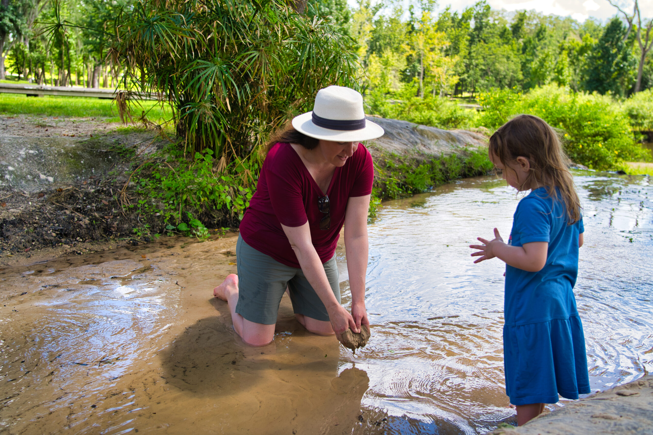 A woman wearing quick-dry KUHL shorts and a sun hat kneels in a shallow creek, scooping up wet sand while a young girl in a blue dress watches nearby. The scene captures a playful, nature-filled moment, emphasizing the shorts' comfort, durability, and ability to dry quickly during outdoor family adventures.