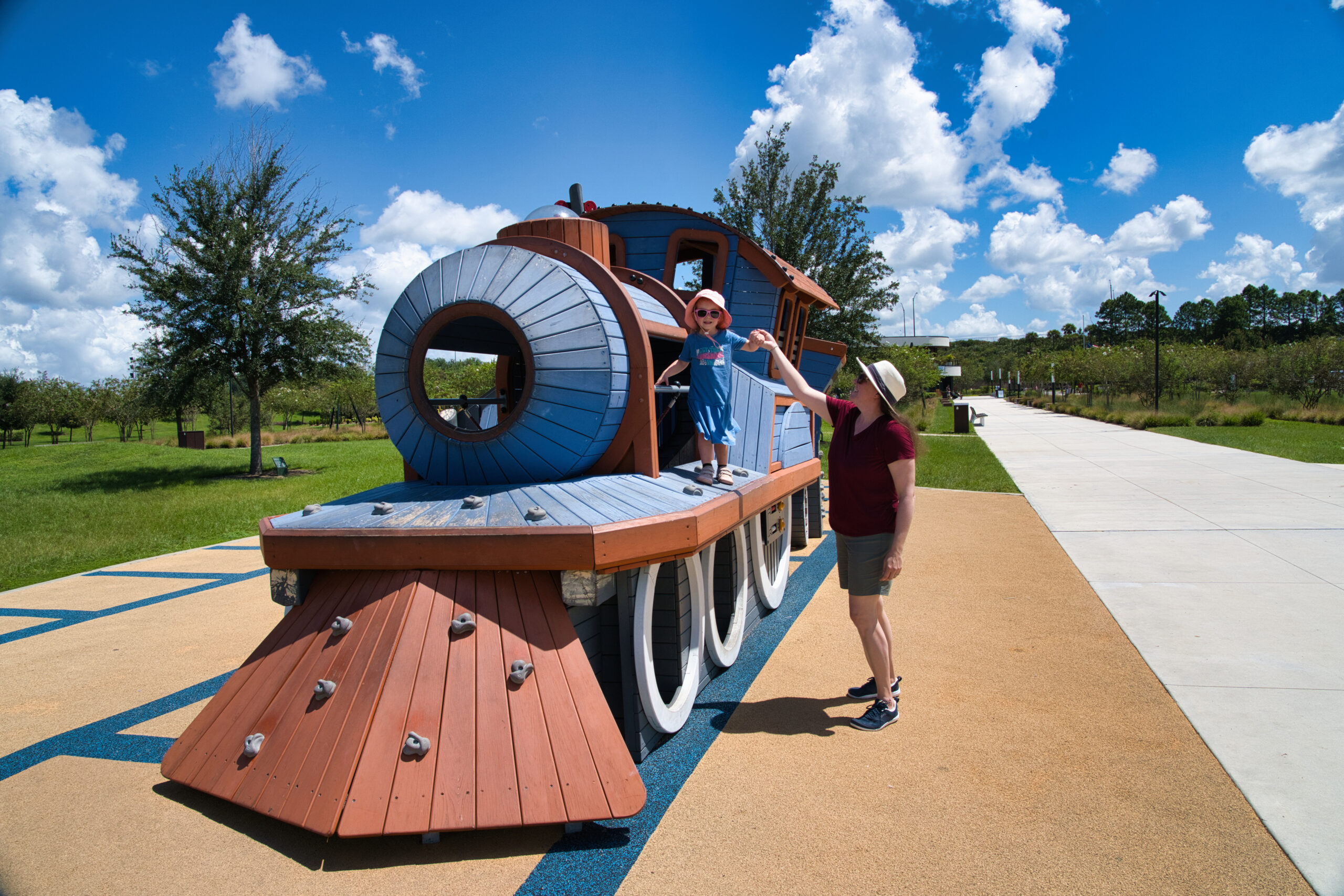 A woman wearing KUHL shorts and a sun hat helps a young girl balance on a colorful wooden train structure at a playground. The playful outdoor setting highlights the shorts' comfort and ease of movement, ideal for active days with kids.
